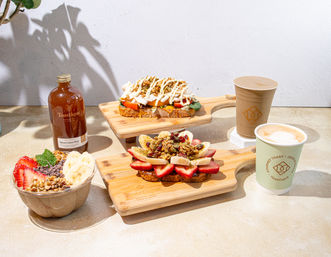 Bright cafe breakfast scene with two open-faced toasts on wooden boards—one topped with sliced strawberries, banana and granola, the other with tomatoes, greens and a creamy drizzle—accompanied by a smoothie bowl, bottled juice and two lattes.