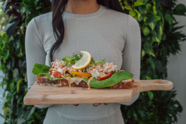 Open-faced crab sandwich on toasted sourdough with melted cheese, tomato, butter lettuce, microgreens and lemon slice on a wooden serving board held against a leafy cafe backdrop, fresh brunch vibe.