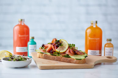 Open-faced smoked salmon toast on rustic sourdough topped with cucumber, microgreens and a lemon slice on a wooden board, with colorful bottled juices and a small bowl of salad blurred in the background — bright healthy brunch scene.