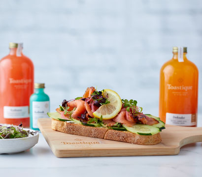 Open-faced smoked salmon toast on rustic sourdough topped with cucumber, microgreens and a lemon slice on a wooden board, with colorful bottled juices and a small bowl of salad blurred in the background — bright healthy brunch scene.