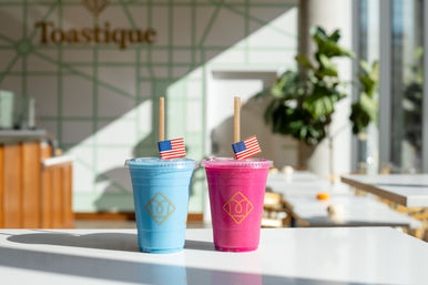 Two colorful fruit smoothies — one blue, one pink — in clear cups with paper straws and tiny American flags, sitting on a sunlit white table in a modern cafe interior with plants and geometric tiled wall.