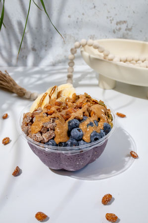 Purple acai smoothie bowl topped with fresh blueberries, banana slices, crunchy granola, candied nuts and nut butter drizzle on a white tabletop