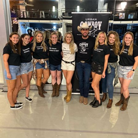 Smiling bachelorette group with cowboy-hat instructor posing in a country line-dancing studio, all wearing casual tees, denim shorts and cowboy boots.