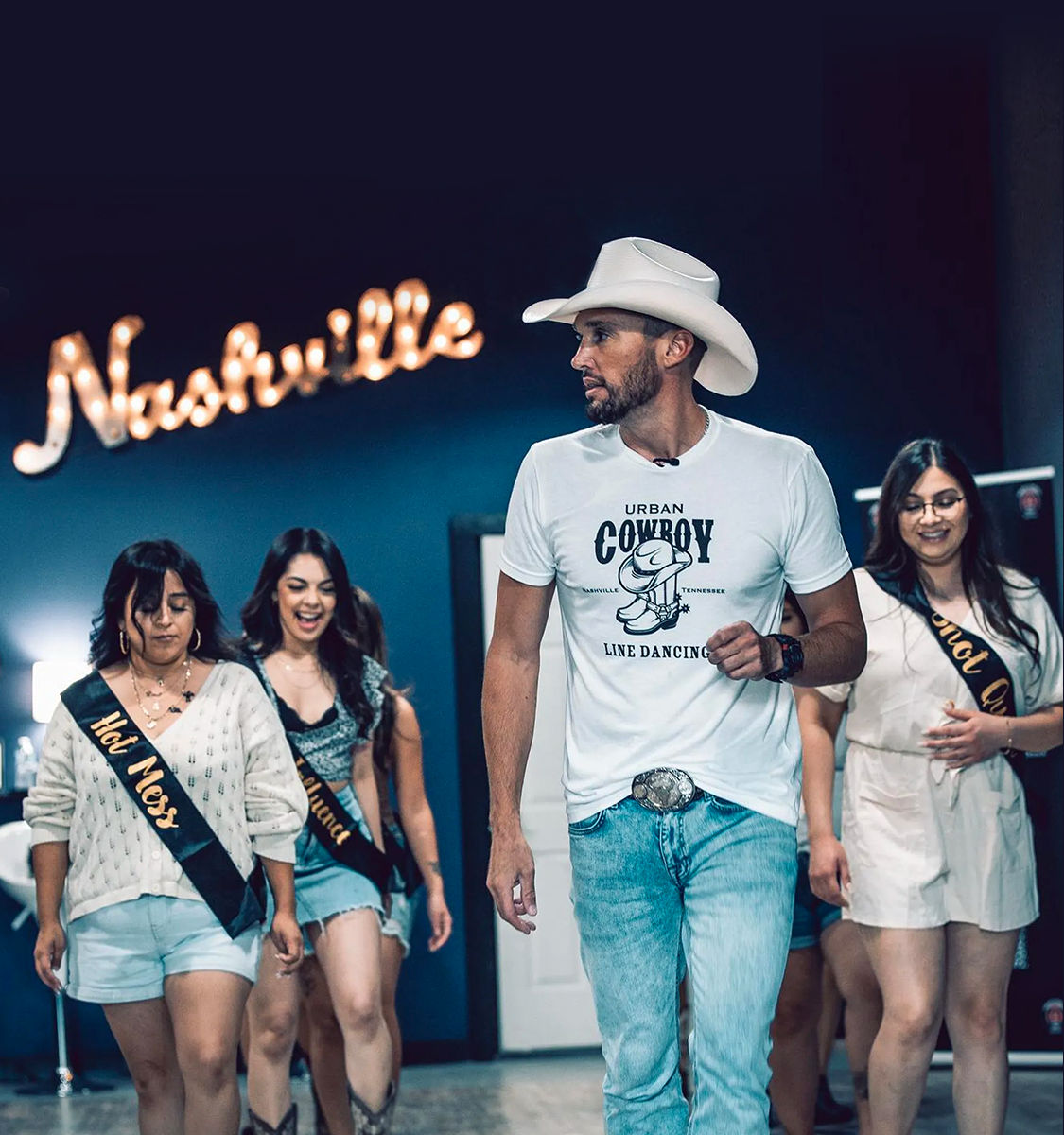 Man in a white cowboy hat leading a Nashville line-dancing class in a studio, neon Nashville sign in the background, wearing a white tee and jeans while three women in sashes follow behind.