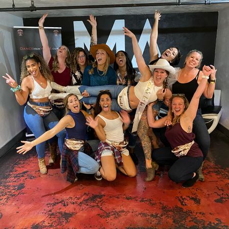 Lively bachelorette party in an indoor dance studio — a dozen women cheer and pose as friends lift the bride-to-be in a sash, veil and cowboy hat; casual outfits, boots and fanny packs, playful celebration.