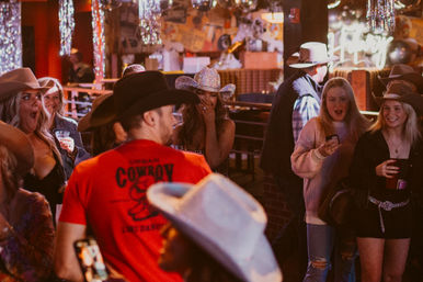 Lively country bar scene with a crowd in cowboy hats, patrons holding drinks and phones, and surprised reactions under neon lights and disco decorations