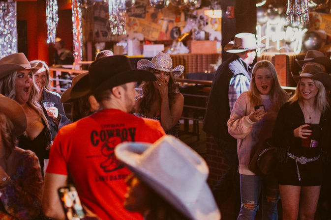 Lively country bar scene with a crowd in cowboy hats, patrons holding drinks and phones, and surprised reactions under neon lights and disco decorations