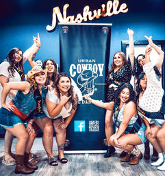 Laughing group of women in cowboy boots and sashes striking playful poses in front of a cowboy-themed banner under a neon "Nashville" sign