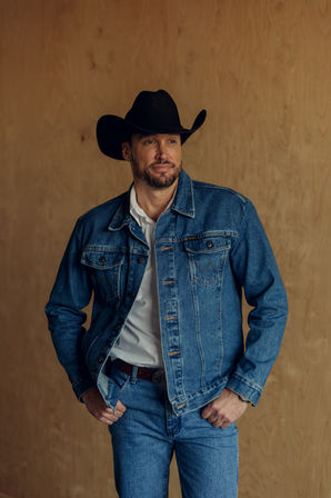 Western-style portrait of a man wearing a black cowboy hat, white shirt, blue denim jacket and jeans with a decorative belt buckle, posing against a light wood backdrop.