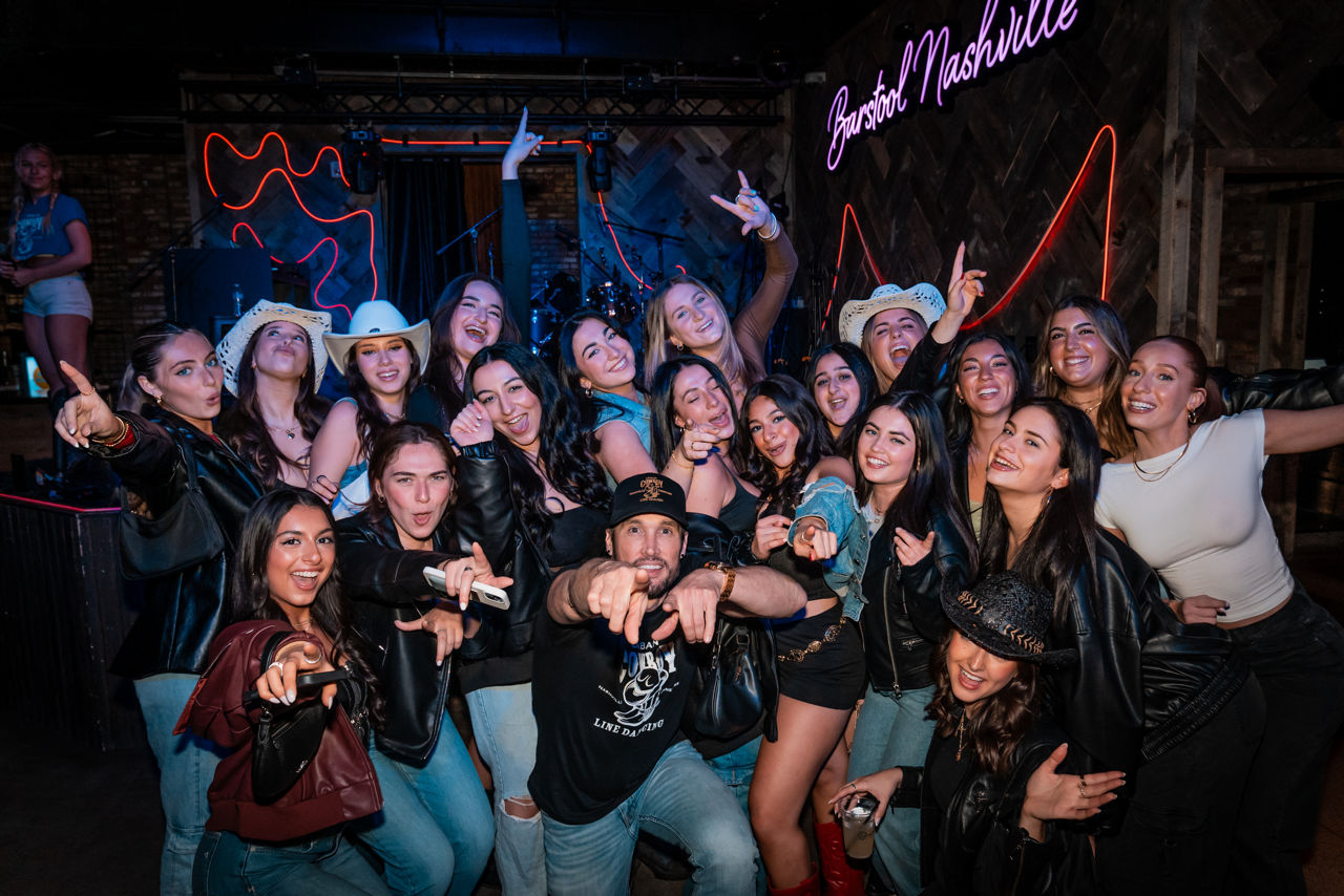 Large group of young adults in cowboy hats and leather jackets posing and pointing at the camera in a neon-lit Nashville bar with a stage in the background