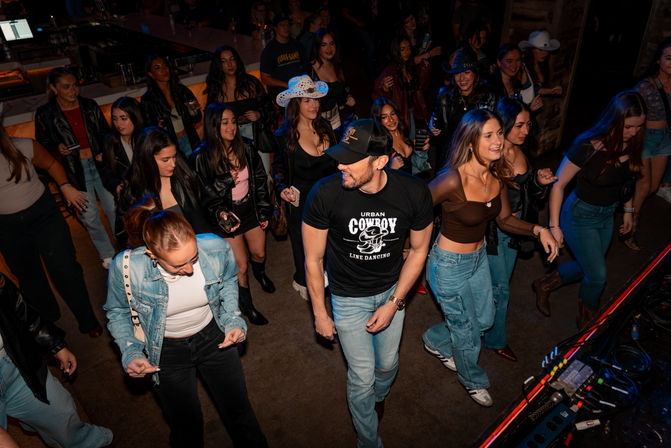 Crowded indoor dance floor at a country‑themed bar with people line dancing and smiling — man in an 'Urban Cowboy Line Dancing' tee, women in denim and cowboy hats, bar and DJ gear visible.