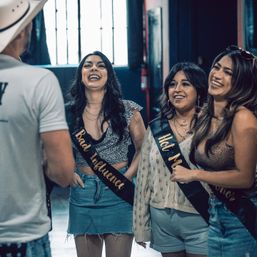 Three laughing women in casual summer outfits wearing novelty sashes ('Bad Influence', 'Hot Mess') chatting with a man in a white hat inside an urban indoor event venue.