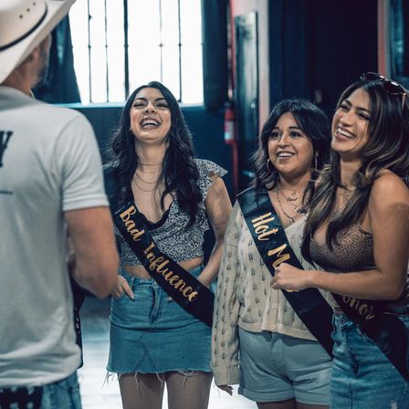 Three laughing women in casual summer outfits wearing novelty sashes ('Bad Influence', 'Hot Mess') chatting with a man in a white hat inside an urban indoor event venue.