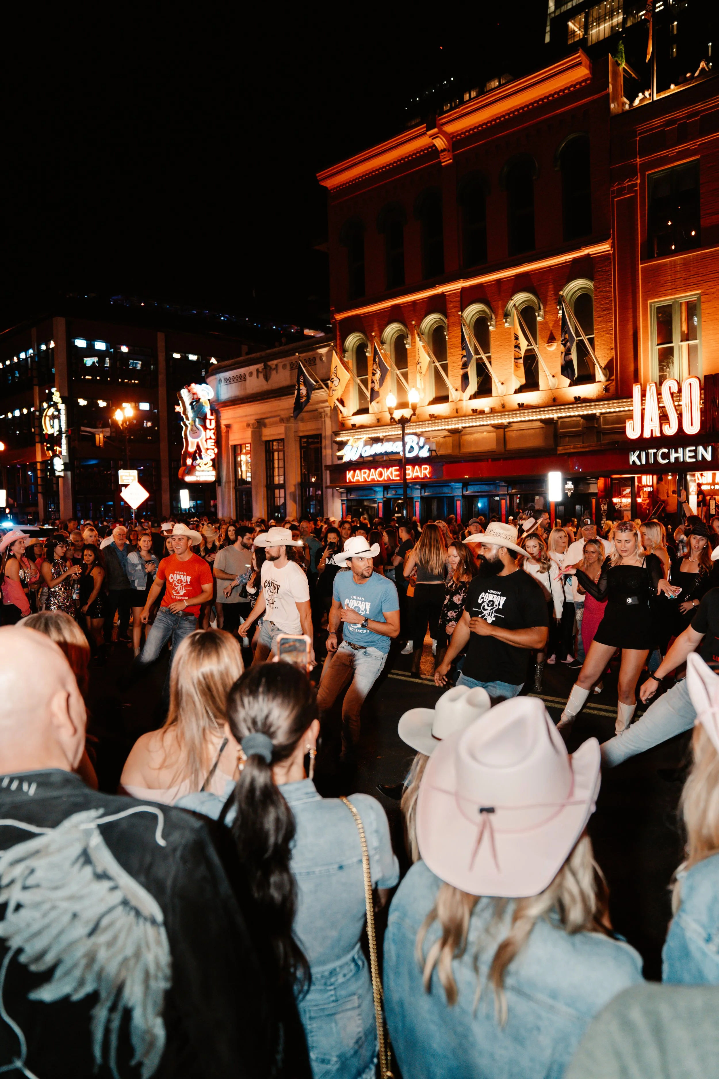 Nighttime downtown street scene with a crowd watching country line dancers in cowboy hats under neon bar signs and glowing historic building facades, lively nightlife atmosphere.
