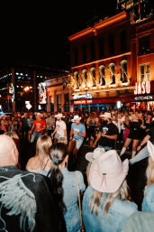 Nighttime downtown street scene with a crowd watching country line dancers in cowboy hats under neon bar signs and glowing historic building facades, lively nightlife atmosphere.