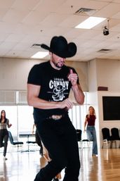 Man in a black cowboy hat and “Urban Cowboy” t‑shirt leading a country line-dance class in a bright indoor dance studio