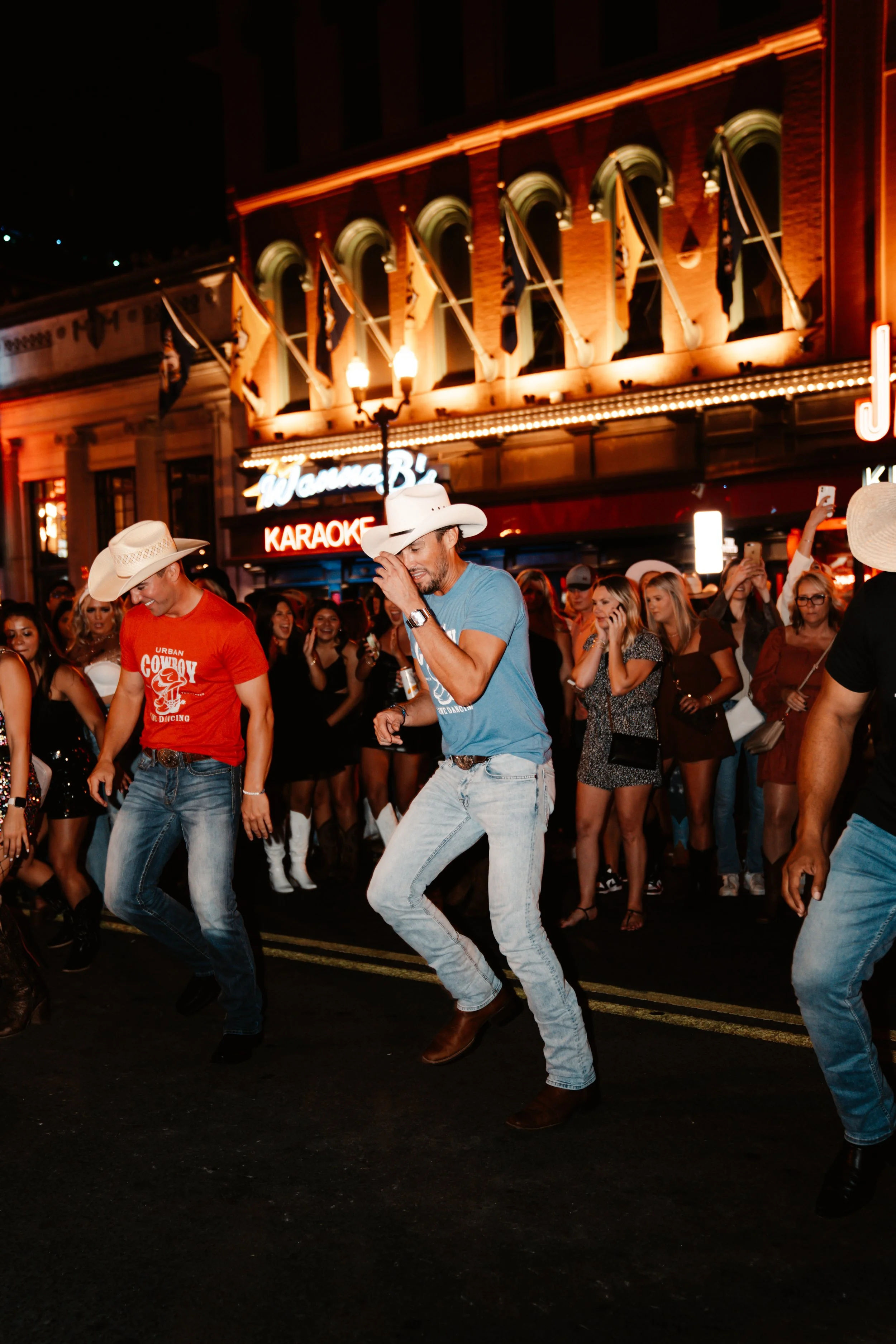Men in cowboy hats and jeans line-dancing on a lively downtown street at night under a glowing marquee and neon karaoke sign, surrounded by a cheering crowd.