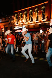 Men in cowboy hats and jeans line-dancing on a lively downtown street at night under a glowing marquee and neon karaoke sign, surrounded by a cheering crowd.