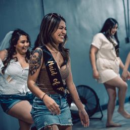 Laughing woman with tattoo wearing a "Maid of Honor" sash and denim skirt leading friends indoors at a casual bachelorette party, bride in a veil behind her.