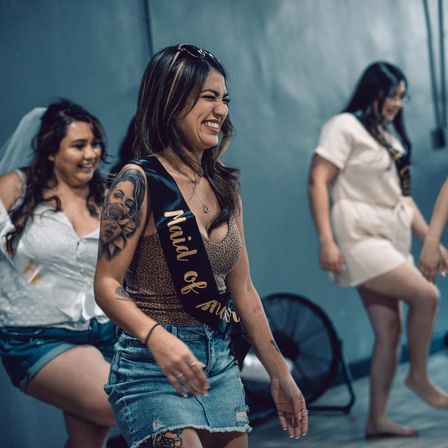 Laughing woman with tattoo wearing a "Maid of Honor" sash and denim skirt leading friends indoors at a casual bachelorette party, bride in a veil behind her.