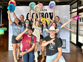 Group of smiling young adults in Nashville-themed t-shirts and colorful cowboy hats posing in a lively indoor photo booth, two men kneel front while others cheer and toss pastel hats