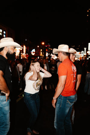 Young woman laughing with two men in cowboy hats on a lively downtown neon-lit street at night, crowd and colorful signs in the background