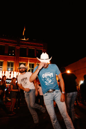 Two men in cowboy hats and jeans line-dancing on a neon-lit downtown street at night, lively urban nightlife with belt buckles and country-style energy.