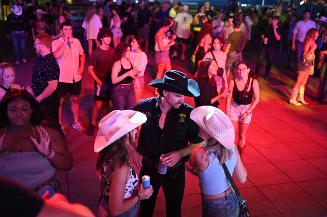 Lively outdoor nightlife scene under pink and purple lights: a man in a black cowboy hat chats with two women in white cowboy hats while a crowd mingles and dances with drinks on a country-music dance floor.