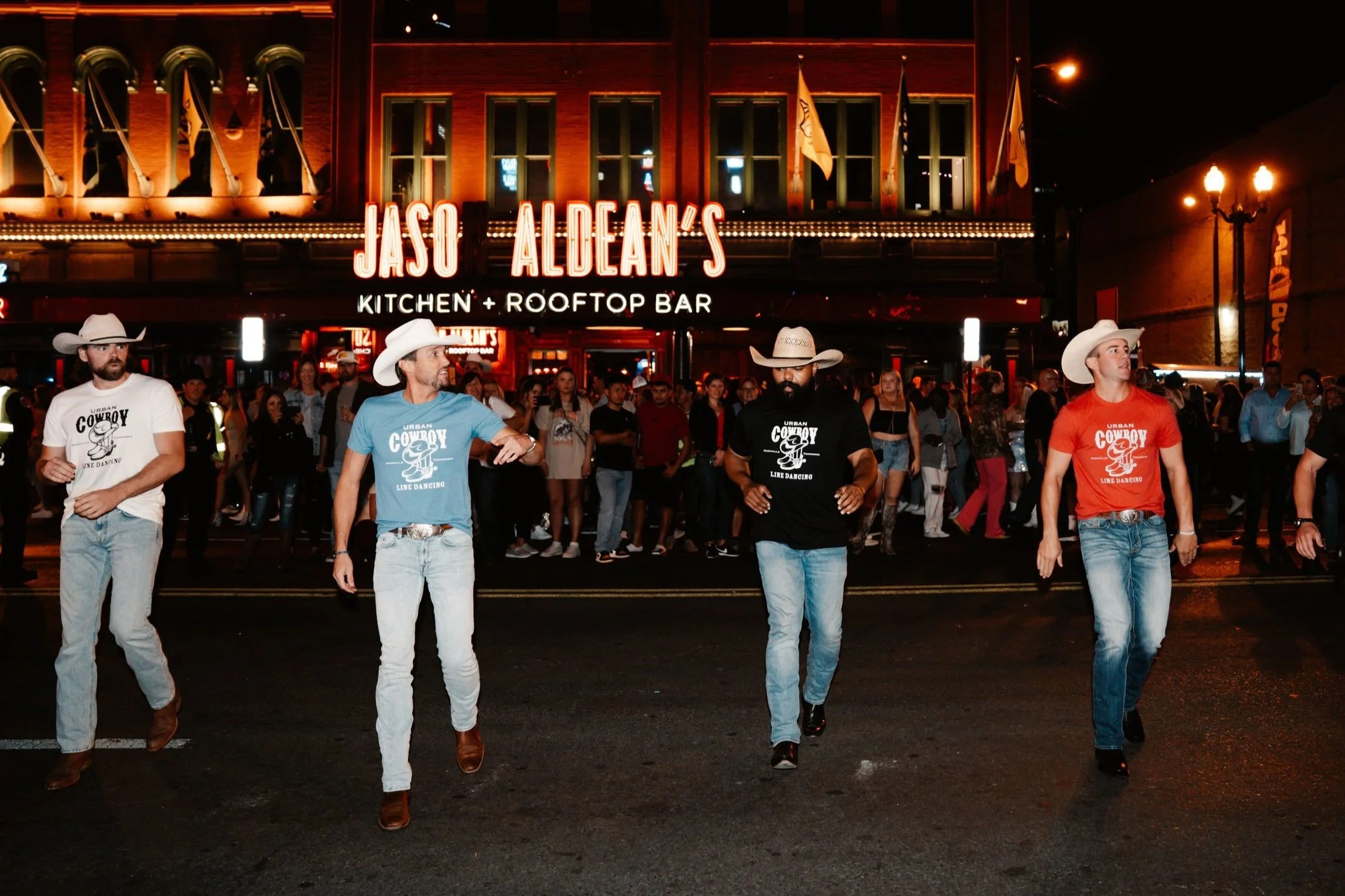 Four cowboy-hat-wearing men line dancing on a downtown street at night in front of a neon-lit rooftop bar and marquee, crowds watching under warm lights.