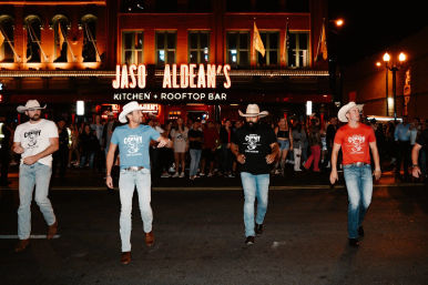 Four cowboy-hat-wearing men line dancing on a downtown street at night in front of a neon-lit rooftop bar and marquee, crowds watching under warm lights.