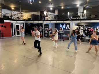 Group country line-dance class in a spacious indoor dance studio, instructor in a cowboy hat leading dancers in boots, hats and casual outfits beneath a mezzanine and hanging lights.