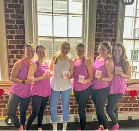 Six smiling women in matching pink workout tanks and leggings pose with drinks inside a sunlit exposed-brick fitness studio with large windows.