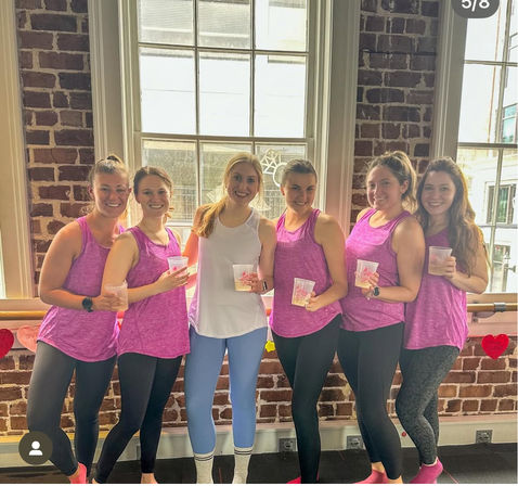 Six smiling women in matching pink workout tanks and leggings pose with drinks inside a sunlit exposed-brick fitness studio with large windows.