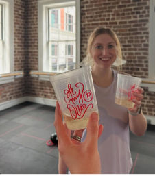Hand holding plastic cup with sparkling drink and stylized red script logo, toasting a smiling person in a brick‑walled urban fitness studio with large windows and a kettlebell on the floor.