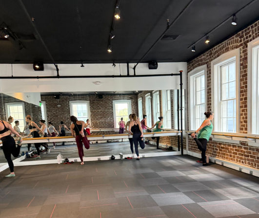 Group barre workout in a brick-walled urban loft-style fitness studio with large windows, mirrored wall, wooden ballet barre and participants stretching and balancing on a rubber-tile floor.