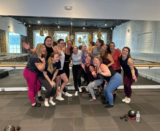 Smiling group of women posing after a barre group fitness class in a bright studio with exposed brick walls, floor-to-ceiling mirrors, ballet barres, kettlebells on the floor and festive gold tassel decorations.