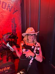 Smiling woman wearing glasses and a white cowboy hat with gold stars in a red-lit bar, holding two beer bottles and a phone beside a decorative horse and western saloon decor.