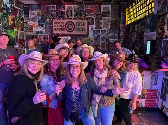 Smiling group of women wearing cowboy hats and star stickers, holding drinks in a lively bar with retro poster-covered walls and neon lighting.