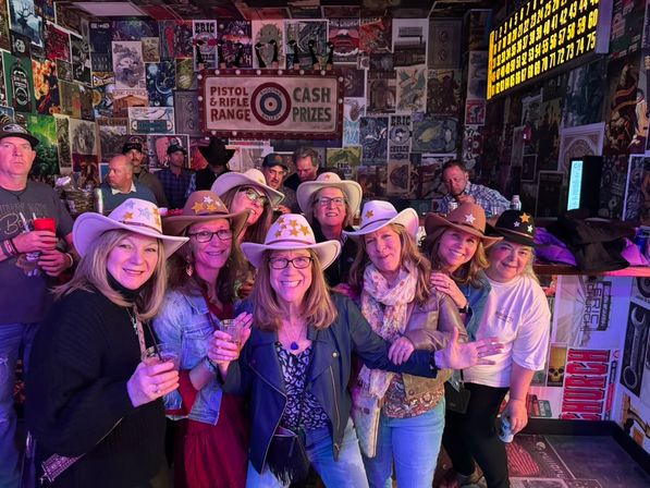 Smiling group of women wearing cowboy hats and star stickers, holding drinks in a lively bar with retro poster-covered walls and neon lighting.