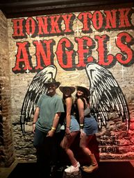 Three people in cowboy hats posing in front of a brick-wall, honky-tonk–style mural with large red lettering and painted angel wings, country bar nightlife vibe