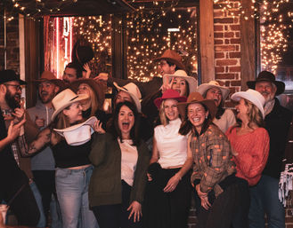 Group of friends wearing cowboy hats, laughing and posing in a rustic brick-walled bar with string lights and a neon sign, lively nighttime party scene.