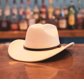 White felt cowboy hat with a black band resting on a polished wooden bar counter, blurred liquor bottles on the back bar in the background