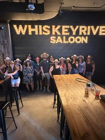 Group of people wearing cowboy hats posing under large illuminated marquee letters on a dark wall inside a rustic saloon, with a long wooden table and drinks in the foreground.