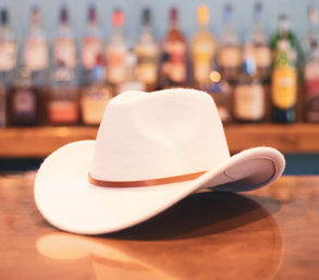 White felt cowboy hat with a thin leather band resting on a wooden bar counter, blurred liquor bottles in the background for a saloon-style vibe