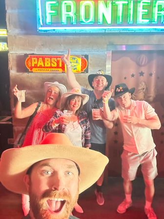 Group selfie of five friends wearing cowboy hats at a western-themed bar, holding drinks and posing happily under neon signs for a lively country-night outing