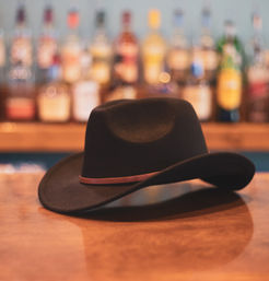 Black felt cowboy hat with a brown leather band resting on a polished wooden bar counter, blurred bottles and warm bokeh lights on the back shelf evoking a cozy western saloon.