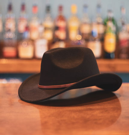 Black felt cowboy hat with a brown leather band resting on a polished wooden bar counter, blurred bottles and warm bokeh lights on the back shelf evoking a cozy western saloon.