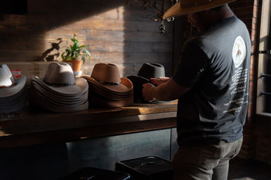 Sunlit rustic hat shop counter with stacks of tan and gray felt cowboy hats and a customer trying on a black hat