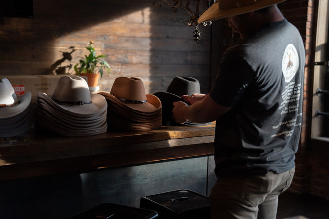 Sunlit rustic hat shop counter with stacks of tan and gray felt cowboy hats and a customer trying on a black hat