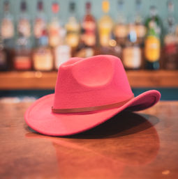 Bright pink felt cowboy hat with a slim leather band resting on a polished wooden bar counter, blurred liquor bottles in the background.